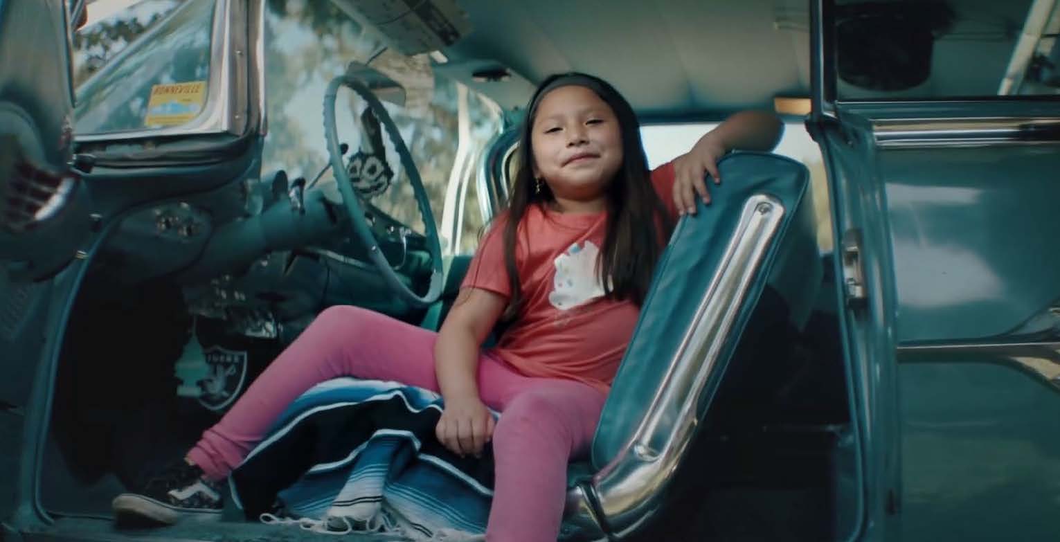 still of a smiling young girl dressed in bright clothing and vans trainers, sat confidently in the driver's seat of a teal car, viewed from a low perspective through the open driver's side door